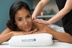 Woman with dark hair smiles at the camera as she receives a deep tissue massage on a table covered in white spa towels