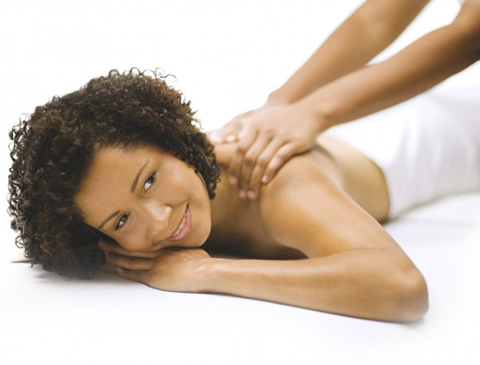 A pair of hands massaging the shoulders of a smiling woman with curly hair while she lays on her stomach